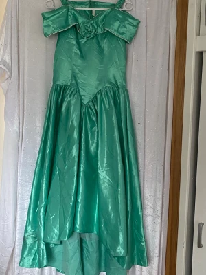 Turkos balklänning  - Turkos balklänning i glansigt material med axelband och dekorativ ros framtill. Klänningen har pärldetaljer längs överdelen och en asymmetrisk kjol. Perfekt för dig som vill sticka ut med en unik look.