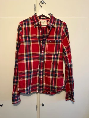 Rutig röd skjorta Abercrombie & Fitch - Snygg rutig skjorta från Abercrombie & Fitch i rött, blått och gult. Klassisk krage, knappstängning framtill och en bröstficka med broderad logga. Tillverkad i mjuk bomull och har långa ärmar. Perfekt för en avslappnad men stilsäker look.