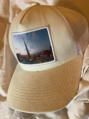 Beige keps med tryck - Snygg beige keps med vit mesh på sidorna och ett stort tryck framtill som föreställer Dubais skyline med Burj Khalifa. Kepsen har böjd skärm och justerbar baksida. Perfekt för dig som gillar streetstyle och unika motiv.