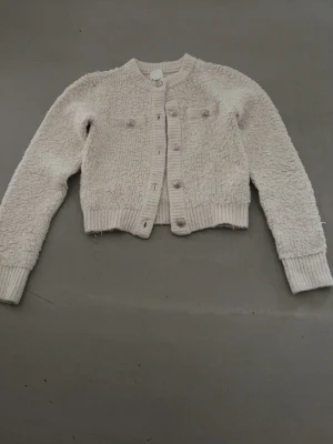 Krämvit teddykofta med knappar - Mysig krämvit teddykofta med rund hals och knäppning framtill. Koftan har ribbade muddar vid ärmslut och nederkant samt två dekorativa bröstfickor. Perfekt för lager-på-lager och en chill look.