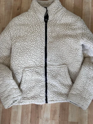 Beige teddyjacka från Brave Soul - Mysig beige teddyjacka från Brave Soul med hög krage och dragkedja framtill. Jackan har två stora fickor och är gjord i fluffigt teddy-material som håller dig varm. Perfekt för kyliga dagar och ger en trendig look.