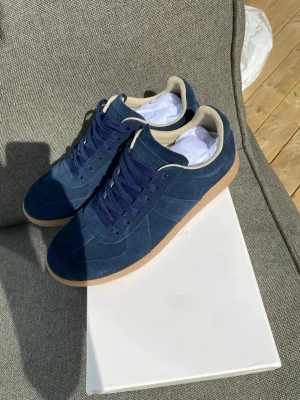 Blå sneakers Maison Margiela - Maison Margiela sneakers i djupblå mocka med beige detaljer och gummisula. Klassisk låg modell med blå skosnören och stilren design. Perfekt för dig som gillar exklusiva och trendiga sneakers. Aldrig använda som man kan se på bilderna. Storlek 44, skickas inom en dag, tveka inte på att höra av dig!/Noah🙂