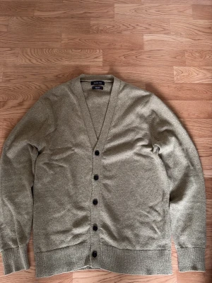 Olivgrön cardigan från Massimo Dutti - Olivgrön cardigan från Massimo Dutti med v-ringning och svarta knappar framtill. Stickad i mjukt material och har ribbade muddar vid ärmslut och nederkant. Perfekt för lager-på-lager och enkel att matcha med olika outfits.