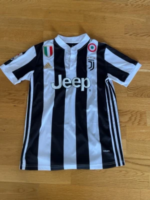 Juventus matchtröja svartvit Adidas - Juventus officiella matchtröja från Adidas i svartvitt randigt med klubbmärke, Jeep-logga och italienska flaggan på bröstet. Tröjan har korta ärmar, klassisk krage och är tillverkad i lätt funktionsmaterial perfekt för fotboll. Hör av er för fler bilder. 