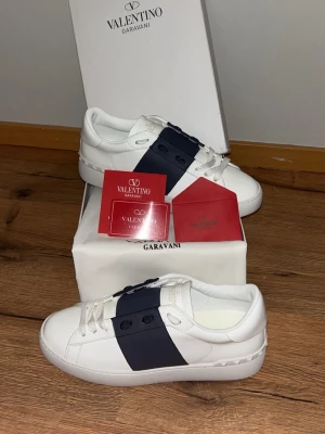 Valentino Garavani vita sneakers - Snygga vita sneakers från Valentino Garavani med en bred mörkblå rand på sidan och vita skosnören. Skorna har en klassisk låg profil och är tillverkade i läder med subtila nitar på hälen. Perfekt för dig som vill ha en stilren och exklusiv look.