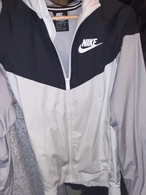 Nike vindjacka svart och grå  - Nike vindjacka i loose fit med dragkedja. Jackan är svart upptill och ljusgrå nedtill, med vit Nike-logga på bröstet. Tillverkad i lätt, vindtätt material och har meshfoder för extra komfort. Perfekt för en sportig och avslappnad stil.