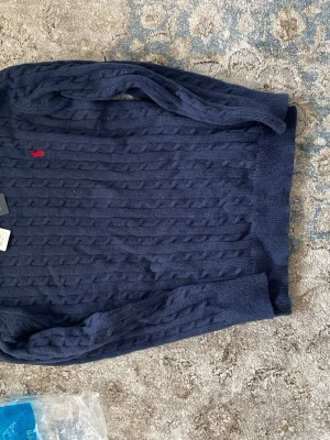 Polo Ralph Lauren tröja - Mörkblå kabelstickad tröja från Polo Ralph Lauren med rund halsringning och den klassiska röda logotypen broderad på bröstet. Tröjan har ribbade muddar vid ärmslut och nederkant. Perfekt för dig som gillar tidlös och stilren design.