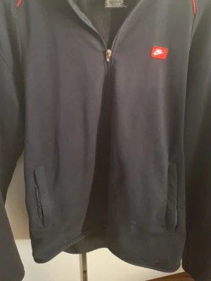 Svart Nike hoodie med dragkedja - Cool svart hoodie från Nike med dragkedja framtill och två fickor. Diskret röd Nike-logga på bröstet och röda detaljer vid axlarna. Tillverkad i mjukt material som är perfekt för en avslappnad look. Passar dig som gillar sportig stil.