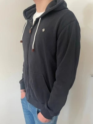 Morris hoddie  - En svart Morris hoodie med dragkedja, vit broderad logga på bröstet och vita snören med bruna detaljer. Hoodien har två stora fickor framtill och ribbade muddar vid ärmslut och nederkant. Perfekt för en avslappnad och cool look.