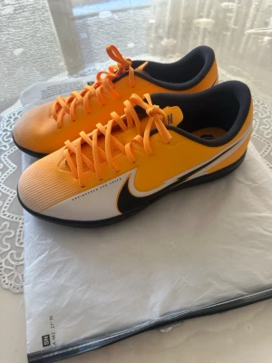 Nike fotbollsskor orange och vit - Nike sneakers i orange och vitt med svarta detaljer och svart yttersula. Skorna har låg profil, klassisk snörning och är tillverkade i syntetmaterial. Stora Nike-loggor på sidan och under sulan ger en sportig och modern look.