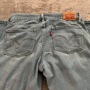 Ljusblå lågmidjade jeans från Levi's - Säljer ett par klassiska blå jeans från Levi's med fem fickor och knappgylf. De har en traditionell passform och är perfekta för en avslappnad stil. 