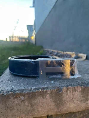 Svart skärp med silverfärgat H-spänne - Snyggt svart bälte med ett stort, silverfärgat H-format spänne. Bältet har vita sömmar längs kanterna och är tillverkat i skinn. Perfekt accessoar för att lyfta din outfit.