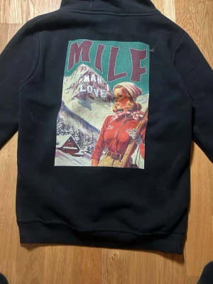 man i love milf hoodie svart - Svart hoodie med stor färgglad man i love milf print på ryggen med text och motiv av en kvinna i skidmiljö. Klassisk huva med dragsko och magficka framtill. Perfekt för dig som gillar streetwear och unika plagg.