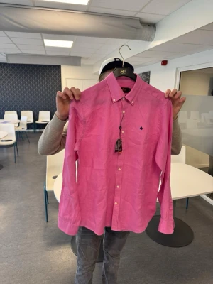 Rosa linneskjorta från morris - Säljer en snygg rosa skjorta från morris i linne. Skjortan har klassisk krage, knappar hela vägen och en liten broderad logga på bröstet. Perfekt för dig som vill ha en stilren och fräsch look med en färg som sticker ut.