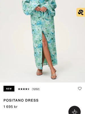 Grön blommig Positano långklänning - Långklänning i Positano-stil med grönt och blått blommönster. Klänningen har långa, vida ärmar, v-ringning och knappar framtill samt en hög slits. Perfekt för sommarens alla tillfällen. Helt ny utan användning med lappen kvar 