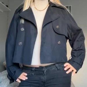 Kort svart trenchcoat - Snygg kort svart trenchcoat med dubbelknäppning och bred krage. Jackan har en croppad passform och klassiska detaljer som stormklaff och stora knappar. Perfekt att slänga över en stickad tröja för en trendig look.