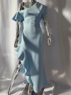 Ljusblå asymmetrisk långklänning - Snygg ljusblå långklänning med asymmetrisk one shoulder-design och volangdetalj längs kjolen. Klänningen har en elegant passform och är tillverkad i ett mjukt, slätt material som ger ett exklusivt intryck. Perfekt för dig som vill sticka ut med en unik och modern look.