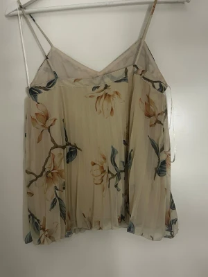 Blommig plisserad linnetopp - Söt linnetopp med tunna axelband och plisserat tyg i ljusbeige med blommigt mönster i grönt och beige. Toppen har en luftig och flowy passform, perfekt för varma dagar. Ingen synlig logga eller märke.