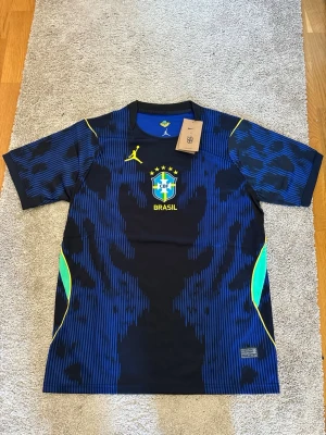 Blå Brasilien fotbollströja Nike - Säljer en blå och svart Brasilien fotbollströja från Nike med Jordan-logga och CBF-emblem på bröstet. Tröjan har gula detaljer och mönstrad design, samt rund halsringning. Perfekt för dig som älskar fotboll och vill stötta Brasilien.