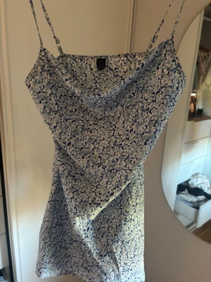 Blommig blå kortklänning med smala band - Söt kortklänning med smala axelband och blått blommönster på ljus botten. Klänningen har en avslappnad passform och lätt draperad urringning. Perfekt för varma dagar och sommarens alla tillfällen.