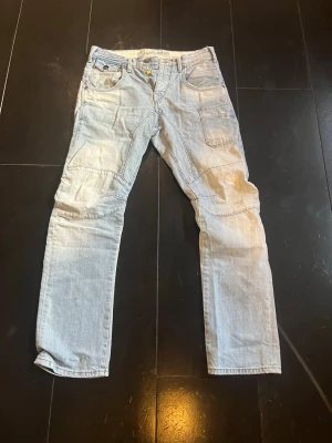 Ljusblå jeans med förstärkta knän - Säljer ett par ljusblå jeans med raka ben och förstärkta knän. Jeansen har flera fickor fram och bak samt detaljer som dubbla sömmar. Materialet är klassisk denim och passformen är avslappnad. Perfekt för dig som gillar en cool och bekväm stil.