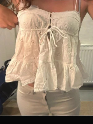 Vit broderad linnetopp med knyt - Söt vit linnetopp med broderade detaljer och volanger. Toppen har knyt framtill och tunna axelband. Perfekt för varma dagar och ger en romantisk känsla till outfiten.