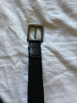 Svart flätat bälte med metallspänne - Snyggt svart flätat bälte med klassiskt metallspänne. Bältet har detaljer i skinn vid spännet och är flexibelt tack vare det flätade materialet. Perfekt för att ge din outfit en clean och stilren touch.