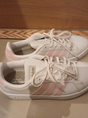 Vita Adidas sneakers med rosa ränder - Snygga vita sneakers från Adidas med klassiska tre rosa ränder på sidan och matchande rosa detalj vid hälen. Skorna har snörning och ovandel i syntetmaterial. Perfekta för en clean och sportig look.