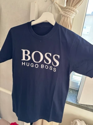 Mörkblå BOSS t-shirts från Hugo Boss - Två mörkblå t-shirts från Hugo Boss med tryck framtill. Ena har stort vitt BOSS-tryck och texten HUGO BOSS under, den andra har ett stilrent BOSS-tryck i beige. Klassisk rund hals och korta ärmar. Perfekta för en clean och enkel stil.