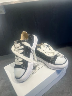 Svarta MMY sneakers med vit sula - Snygga svarta sneakers från Maison Mihara Yasuhiro med vit, chunky sula och kontrasterande vita sömmar. Skorna har breda vita skosnören och en unik, vågig sula som sticker ut. Ovandelen är i canvas och tån är vit. Perfekt för dig som gillar streetwear och statement sneakers.