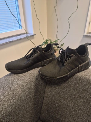 Svarta Adidas NMD sneakers 39 - Säljer ett par svarta Adidas NMD sneakers med klassiska tre ränder på sidan. Skorna har en låg profil, mesh-överdel och gummisula med detaljer på sidan. Perfekta för dig som gillar stilrena och sportiga sneakers.