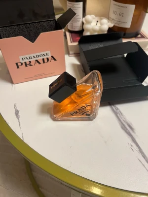 Prada Paradoxe Eau de Parfum 90ml - HELT FULL!!!Prada Paradoxe Eau de Parfum i en unik, triangelformad flaska med svart kork och transparent glas som visar den orangefärgade parfymvätskan. Medföljer en stilren rosa kartong med tydlig Prada-logga. Perfekt för dig som vill ha en lyxig och modern doft.