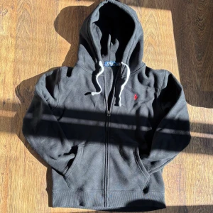 Svart hoodie från Polo Ralph Lauren - Svart hoodie med dragkedja från Polo Ralph Lauren, klassisk design med vit dragsko och röd logotyp på bröstet. Mjuk bomullsmix och rymlig huva gör den perfekt för chill dagar. Fickor framtill och ribbade muddar för extra komfort.