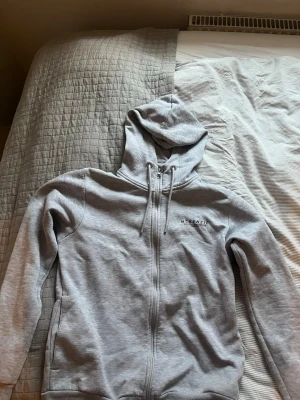 Ljusgrå hoodie från McKenzie - Ljusgrå hoodie från McKenzie med dragkedja framtill och justerbar huva med snören. Tröjan har diskret logga på bröstet och är tillverkad i mjukt bomullsmaterial. Perfekt för en avslappnad och stilren look.