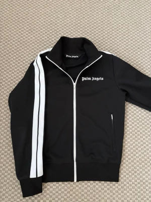 Palm Angels svart zip-tröja (M) - Svart zip-tröja från Palm Angels med vita dubbla ränder längs ärmarna och logotyp på bröstet. Hög krage och ribbade muddar. Materialet är troligtvis polyester, perfekt för en sportig och trendig look.