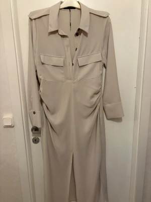 Beige långklänning med fickor från Zara - Stilren beige långklänning från Zara med långa ärmar, stora bröstfickor och klassisk krage. Klänningen har knappar framtill och en slits nedtill för extra rörelsefrihet. Perfekt för dig som gillar minimalistisk och trendig stil.