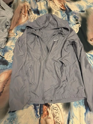 Ljus grå  vindjacka från Nike - Snygg ljuslila vindjacka från Nike med huva och dragkedja framtill. Jackan har två fickor med dragkedja och diskret Nike-logga på bröstet. Tillverkad i lätt och smidigt material, perfekt för blåsiga dagar.