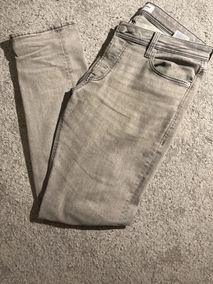 Grå slim fit jeans från Jack & Jones - Snygga grå slim fit jeans från Jack & Jones med klassisk femficksdesign och diskreta bakfickor med broderad detalj. Jeansen har en något tvättad look och är tillverkade i mjuk bomullsdenim med stretch för extra komfort.