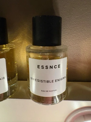 Essnce Irresistible Enigma parfym - En stilren parfymflaska från Essnce med namnet Irresistible Enigma. Flaskan är cylindrisk i klart glas med svart matt kork och vit etikett med minimalistisk design. Innehållet är gyllene och flaskan har en modern look som passar dig som gillar trendiga dofter.