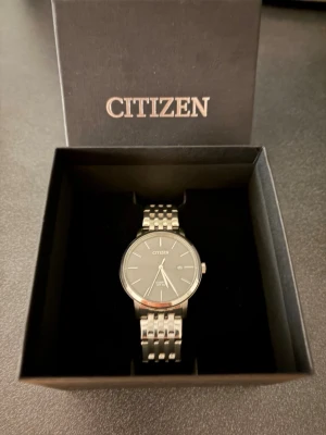 Citizen silverfärgad klocka - Stilren klocka från Citizen med silverfärgat armband i metall och svart urtavla. Klockan har minimalistisk design med datumvisning och klassiskt rund boett. Perfekt accessoar för dig som gillar en clean och tidlös look. Helt ny, aldrig använt så storleken inte passar. Kvitto kan skickas med.