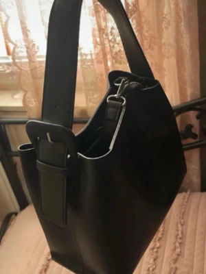 Svart bucket bag med bred axelrem - Stilren svart handväska i bucket-modell med bred justerbar axelrem och silvriga metalldetaljer. Väskan är tillverkad i slätt konstläder och har en minimalistisk design utan synliga loggor. Perfekt för dig som gillar enkel och modern stil.