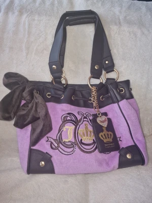 Lila Juicy Couture handväska med detaljer - Snygg lila handväska från Juicy Couture med svarta läderdetaljer, guldfärgade metalldelar och broderade kronor framtill. Väskan har två handtag, dekorativ rosett och en hängande tag med hjärta. Perfekt för dig som gillar statement-accessoarer.