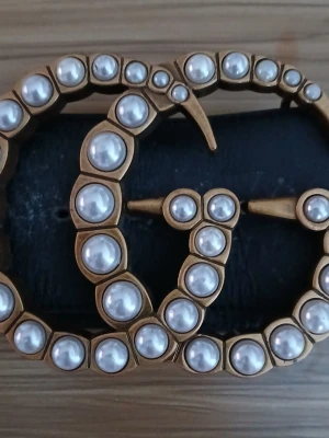 Gucci svart bälte med pärldetaljer - Snyggt svart bälte från Gucci med ikoniskt GG-spänne i guldton och dekorerat med vita pärlor. Bältet är brett och har en lyxig känsla som verkligen sticker ut. Perfekt accessoar för att lyfta vilken outfit som helst.