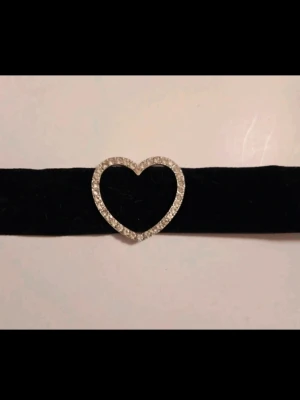 Choker med hjärtformad strass - Brett sammetshalsband i svart med ett glittrande hjärta i mitten. Hjärtat är dekorerat med små stenar. Perfekt accessoar för att lyfta din outfit. Ena sidan har ärjat lite som syns på bild, syns inte när man bär och kan säkert tvättas bort bort Kan även hämtas I Partille  J