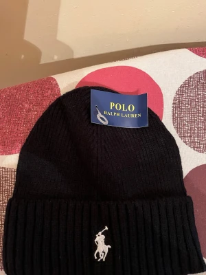 Svart ribbstickad mössa Polo Ralph Lauren - Svart ribbstickad mössa från Polo Ralph Lauren med klassisk uppvikt kant och broderad vit logotyp framtill. Snygg och enkel design som passar till det mesta. Tillverkad i mjukt material för extra komfort.