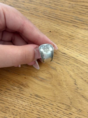 Silverfärgad ring med mönster - Säljer en bred silverfärgad ring med snygga, detaljerade gravyrmönster. Ringen har en vintagekänsla och är tillverkad i metall med ett matt och blankt inslag. Perfekt för dig som gillar unika accessoarer med stil.