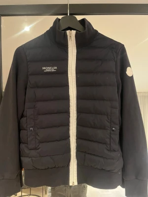 Svart cardigan från Moncler - Svart äkta cardigan från Moncler med vit dragkedja, quiltad front och Moncler-logga på bröstet och ärmen. Jackan har två fickor med knapp och ribbade muddar. 100 % bomull Och 100 % nylon pris kan diskuteras 