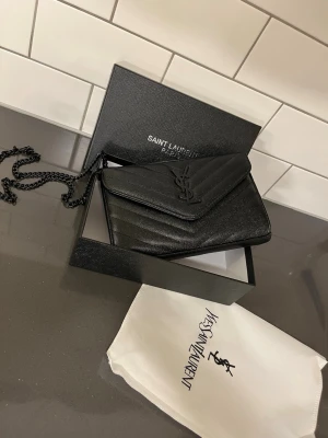 Dam väska - Snygg svart kuvertväska från Saint Laurent i läder med kedjerem och ikoniska YSL-loggan framtill. Väskan har ett elegant kuvertformat lock och diskret struktur i materialet. Perfekt accessoar för att lyfta din outfit.