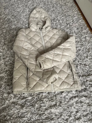 Beige pufferjacka med huva - Snygg beige pufferjacka med huva och dragkedja framtill. Jackan har quiltat mönster och två fickor med dragkedja. Perfekt för kalla dagar och ger en trendig look.