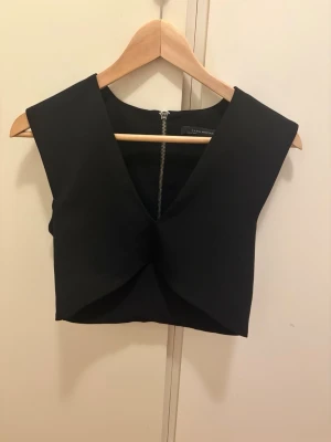 Svart croppad topp från Zara - Stilren svart croppad topp från Zara med djup v-ringning och markerade axlar. Toppen har en dekorativ dragkedja med ring baktill och är ärmlös. Perfekt att styla med höga byxor eller kjol.